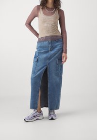 Denim maxi skirt with side slits and cargo pockets, paired with a fitted long-sleeve top in beige and mauve, accentuated by a silver chain.