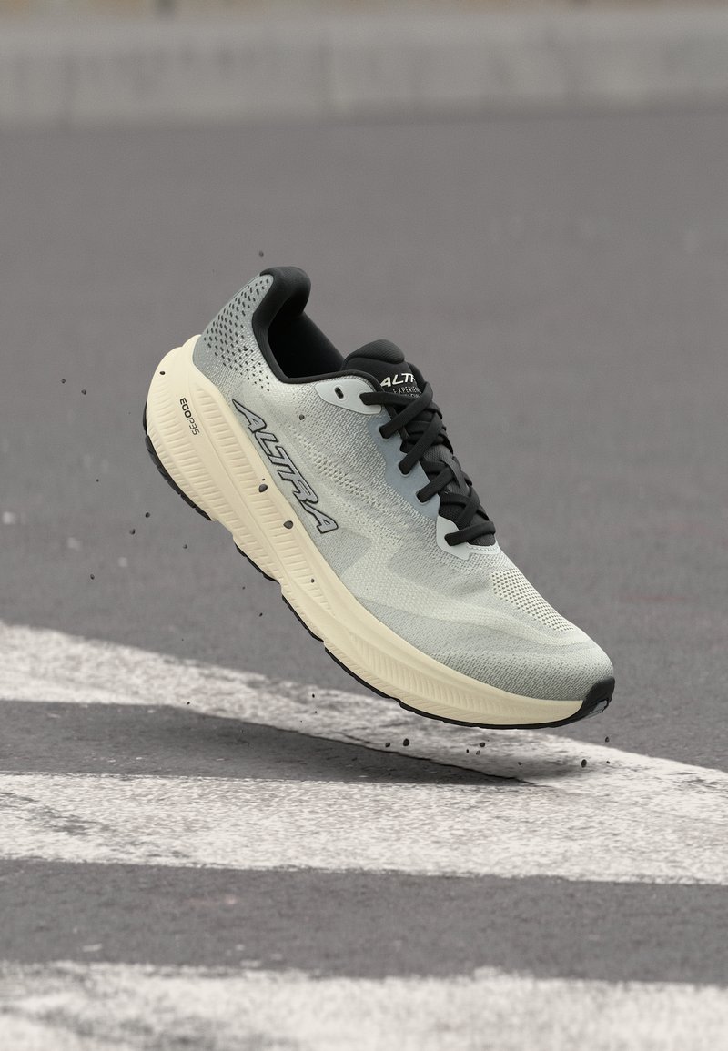 Chaussure de course gris clair et noire avec semelle épaisse crème, lacets noirs et logo "ALTRA", capturée en l'air au-dessus d'une surface en asphalte.
