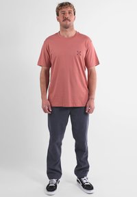 T-shirt rose en coton à manches courtes avec un petit motif noir sur la poitrine, associée à un pantalon en velours côtelé gris foncé et des baskets noires.