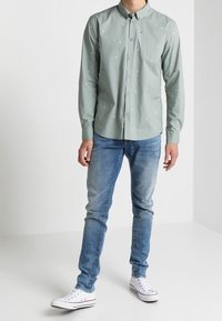 Ljust grön skjorta med knappar och små vita och blå mönster, tillsammans med blå jeans och vita sneakers. Tyget verkar slätt och avslappnat.