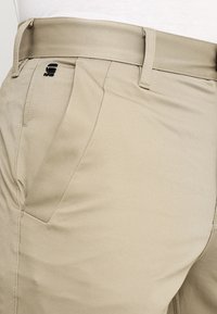 Béžové kalhoty z hladké látky s rovnými předními díly a bočními kapsami, s vyšitým logem značky poblíž pasu.
