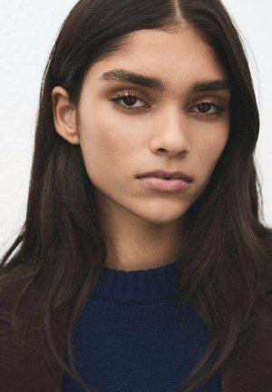 Retrato de una persona con cabello largo y oscuro y maquillaje natural. Lleva un suéter azul marino con un cuello acanalado y una chaqueta oscura.