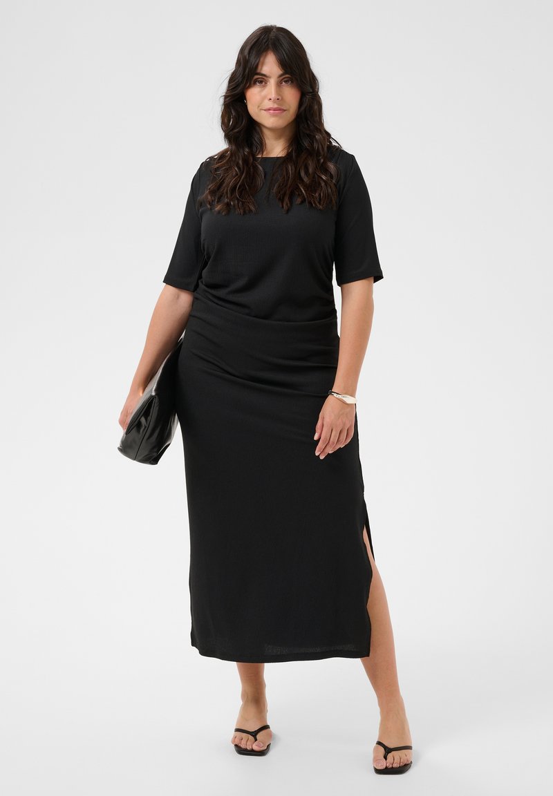 Femme aux longs cheveux foncés portant une robe midi noire avec une fente latérale, tenant une pochette noire, et portant des sandales noires et un bracelet.