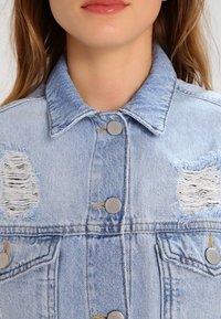 Zbliżenie na osobę w jasnoniebieskiej dżinsowej kurtce z przetarciami i metalowymi guzikami, ukazującą kołnierz i górną część klatki piersiowej.
