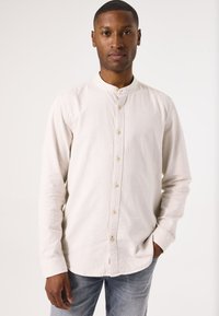 Camisa de manga larga de color beige claro con cuello de banda, cierre frontal de botones y una tela texturizada. Se lleva con jeans de mezclilla claros.