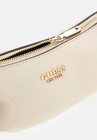 Bustina in pelle sintetica beige con cerniera color oro e logo GUESS. Presenta una texture liscia e una forma curva.