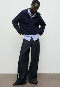 Suéter ancho azul marino con cuello con cremallera, superpuesto a una camisa a rayas azul clara; pantalones vaqueros oscuros de corte holgado con acentos laterales.