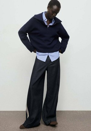 Suéter ancho azul marino con cuello con cremallera, superpuesto a una camisa a rayas azul clara; pantalones vaqueros oscuros de corte holgado con acentos laterales.