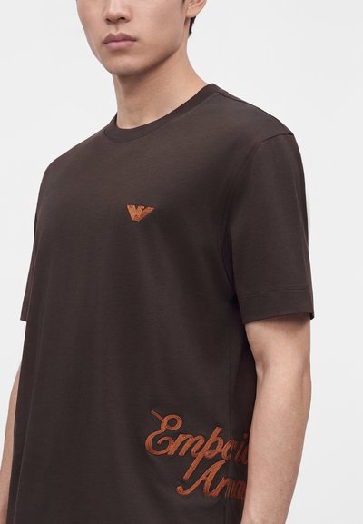 Νέος άνδρας που φοράει ένα σκούρο καφέ T-shirt Emporio Armani με μικρό λογότυπο στο στήθος και μεγάλο κεντητό κείμενο στο πλάι.