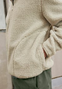 Veste en polaire couleur crème avec une finition texturée, fermeture éclair à l'avant et coupe décontractée. Dispose de poches latérales et de poignets côtelés.