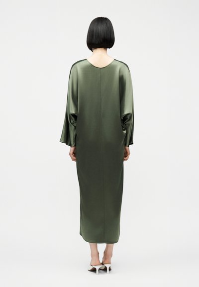 Γυναίκα με κοντά μαύρα μαλλιά, που φοράει ένα μακρύ, φαρδύ σατέν φόρεμα σε ελιά πράσινο χρώμα και λευκές γόβες με ανοιχτή φτέρνα, στέκεται γυρισμένη με την πλάτη.