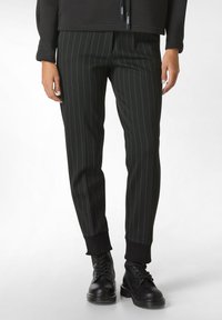 Zwarte pinstriped broek met groene strepen, tapered fit, elastische boorden en zakken, gedragen met zwarte veterschoenen.
