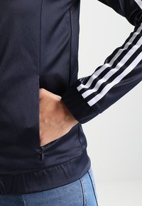 Chaqueta deportiva azul marino con acentos en blanco de tres franjas en las mangas, que cuenta con un bolsillo con cremallera y una textura suave y brillante.