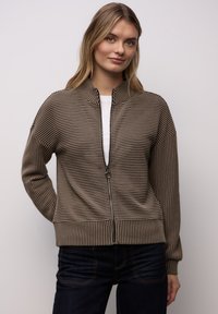 Cardigan marrón a rayas con cremallera y cuello alzado, textura acanalada y hombros caídos, combinado con una camisa blanca debajo.