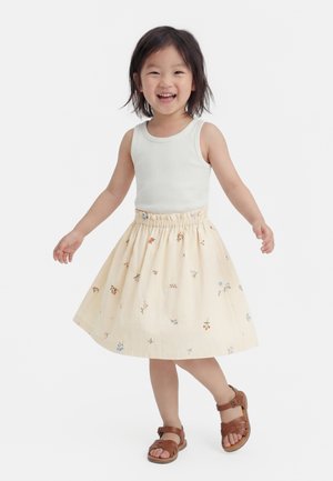 Malé dieťa sa usmieva, nosí biely tielkový top, krémovú kvetinovú sukňu a hnedé sandále, stojí na bielom jednoduchom pozadí.