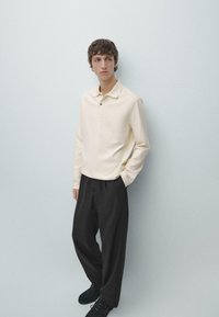 Kremowa koszulka polo z długim rękawem, z kołnierzykiem i dwoma guzikami przy szyi, połączona z czarnymi spodniami o szerokich nogawkach oraz czarnymi butami.