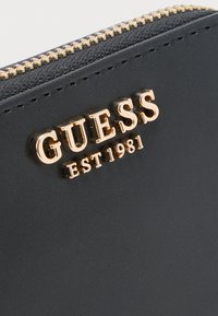 Cartera de cuero negro con cremallera dorada y logo dorado elevado "GUESS EST 1981" en la parte delantera.