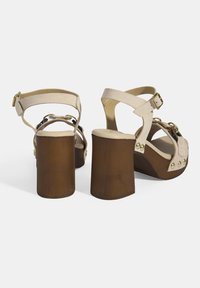Beige Plattformsandalen mit holzähnlichen Absätzen, verstellbaren Riemen, goldfarbenen Hardware und Nietenakzenten im Obermaterial für zusätzliche Details.