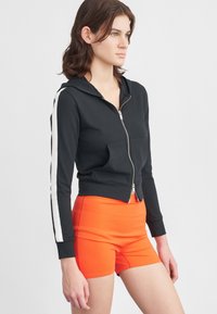 Hoodie preto cropped com riscas laterais brancas, fecho de correr à frente e bolsos duplos. Combinado com calções de cintura alta laranja brilhante. Textura suave.