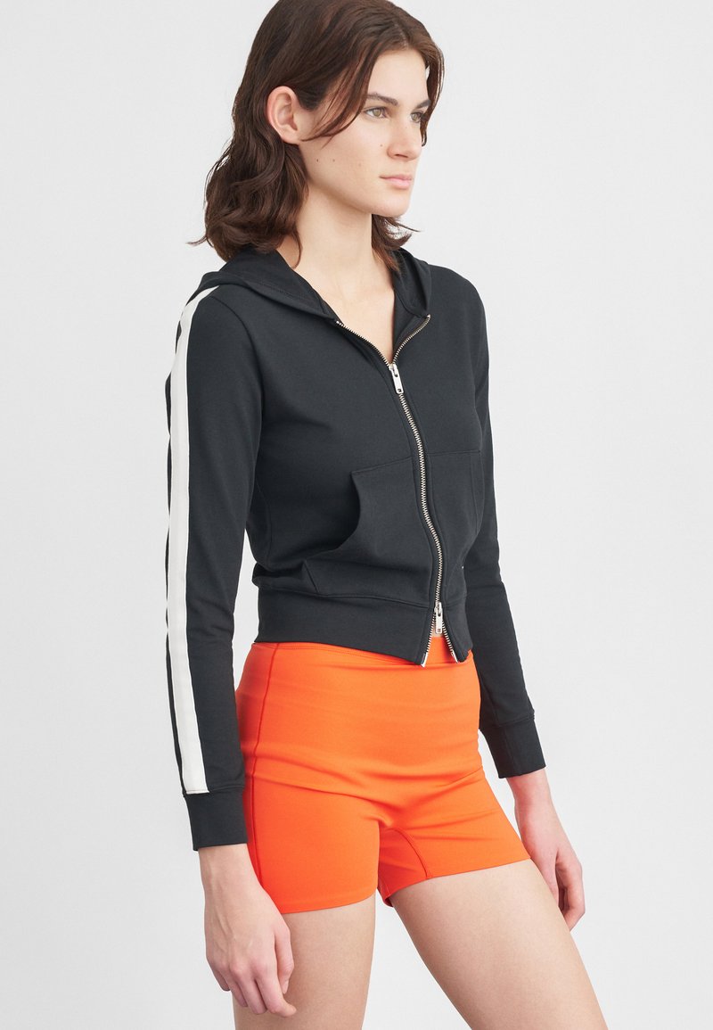 Hoodie preto cropped com riscas laterais brancas, fecho de correr à frente e bolsos duplos. Combinado com calções de cintura alta laranja brilhante. Textura suave.
