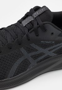 Zapatilla deportiva negra con parte superior de malla y acentos texturizados, un logo de Asics en gris y una suela con patrones. Nombre del modelo: Patriot 13.