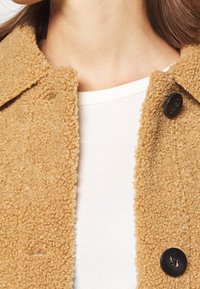 Giacca in shearling color beige con una superficie testurizzata e soffice. Presenta un colletto a revers e due bottoni scuri sul davanti. Sotto, una maglietta bianca.