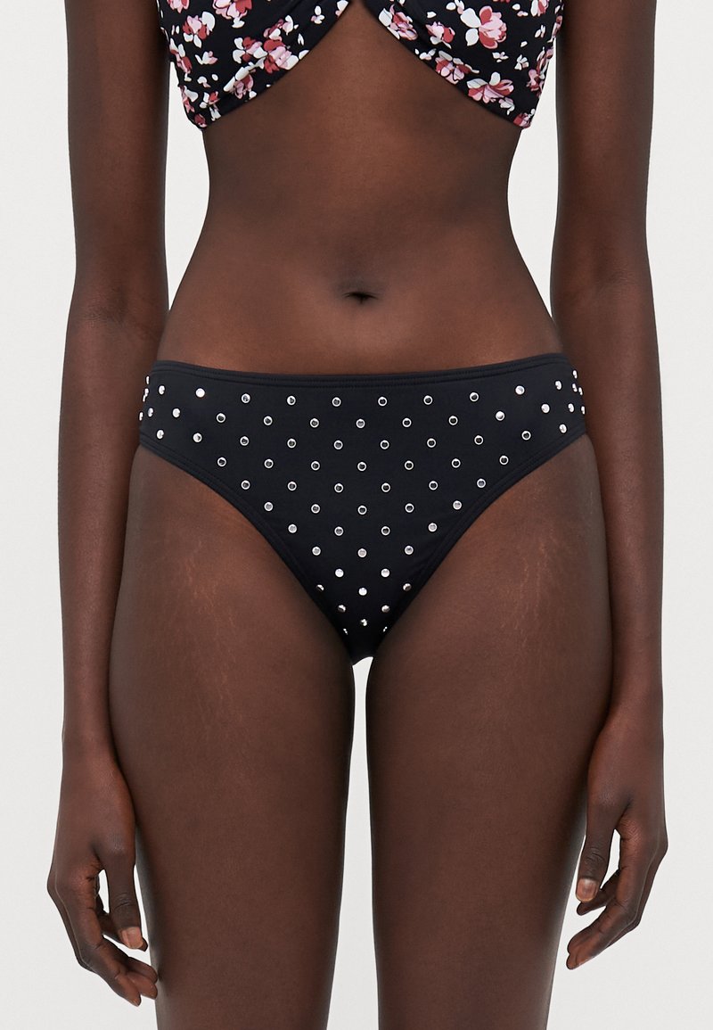 Bas de bikini noir avec des clous en argent circulaires, taille plate et coupe classique. Texture lisse avec un design ajusté.