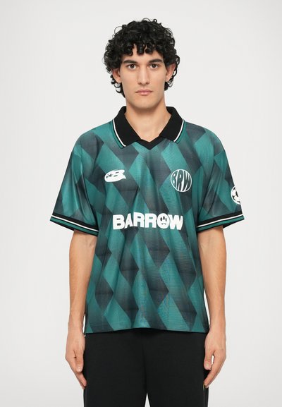 Maillot de sport à motifs verts et noirs, avec un col, des manches courtes et des logos sur le devant. Le design en losanges texturé ajoute de la profondeur.