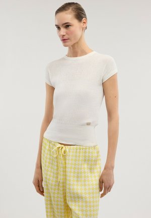 Frau trägt ein weißes, geripptes Kurzarmoberteil und gelb-weiß karierte Jogginghosen mit Kordelzug, steht vor einem einfarbigen Hintergrund.