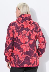 Žena s krátkými blond vlasy, oblečená v kapuci s květinovým vzorem v purpurové a červené barvě, viděná zezadu.