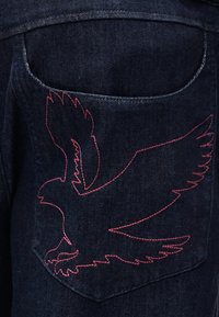 Πίσω τσέπη από σκούρο τζιν με ροζ κέντημα που σχηματίζει περίγραμμα πουλιού σε πτήση, με απλωμένα φτερά και ανοιχτό ράμφος.