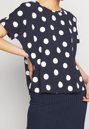 Kobieta ubrana w luźną granatową bluzkę w duże białe grochy oraz dopasowaną prążkowaną ciemną spódnicę.