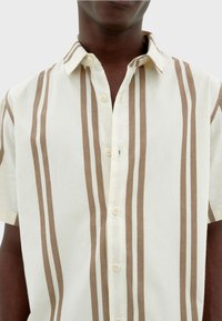 Camisa de botones de manga corta en beige con rayas verticales marrones. Confeccionada en tela ligera, presenta un cuello con solapa y un acabado texturizado.