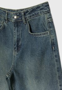 Vaqueros de denim en un tono azul desgastado, con cierre de botón, trabillas para el cinturón y bolsillos frontales con costuras amarillas en contraste.