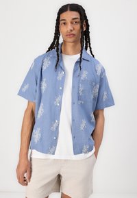 Chemise à manches courtes bleu clair avec broderies florales blanches, une poche à rabat et un col décontracté, portée sur un T-shirt blanc.
