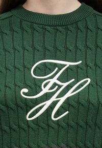 Roheline soonikkoes kampsun, mille keskmes on silmapaistev valge tikandiga monogramm. Tekstuuriga muster lisab visuaalset huvi.