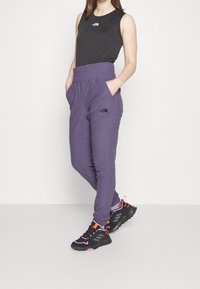 Lilas Jogginghosen mit elastischem Taillenbund, seitlichen Taschen und schwarzem Logo-Detail am Oberschenkel, kombiniert mit einem schwarzen ärmellosen Top und sportlichen Sneakern.