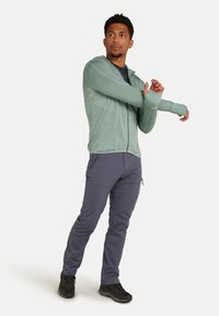 Felpa con zip verde chiaro con un motivo testurizzato, abbinata a pantaloni grigi scuri aderenti e scarpe nere. Il modello sta allungando le braccia.