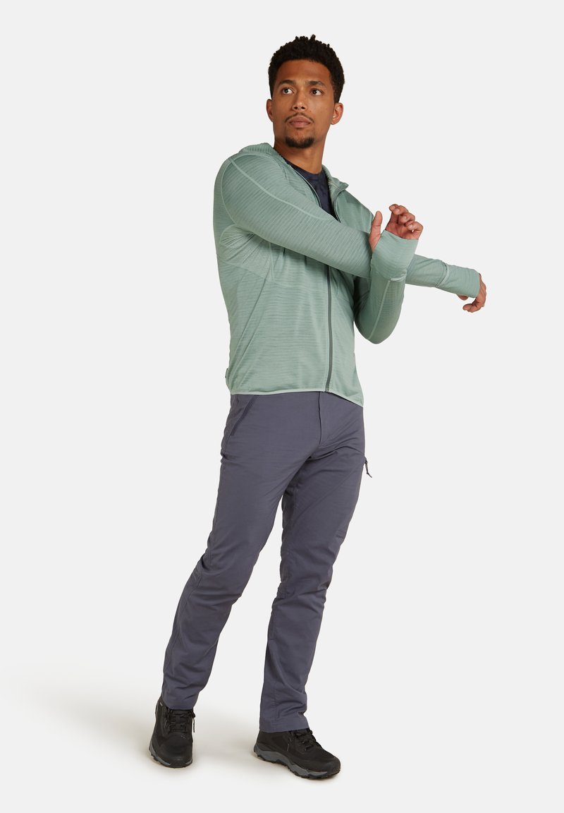 Felpa con zip verde chiaro con un motivo testurizzato, abbinata a pantaloni grigi scuri aderenti e scarpe nere. Il modello sta allungando le braccia.
