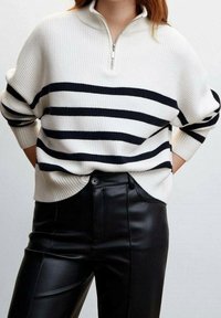 Weißer und marineblau gestreifter Strickpullover mit einem Viertel-Reißverschluss am Kragen; getragen über schwarzen Kunstlederhosen. Der Pullover hat eine lockere, lässige Passform.