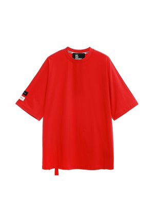 Joven mujer de pie, vistiendo una camiseta roja holgada, pantalones cargo blancos con detalles de texto y botas blancas con cordones sobre un fondo blanco liso.