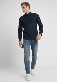 Navyblaues Hemd mit langen Ärmeln und slim fit, kombiniert mit hellblauen Jeans und weißen Sneakern vor einem neutralen Hintergrund.