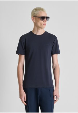 Joven con cabello corto y rubio, lleva gafas de sol negras, una camiseta lisa de color azul marino oscuro y pantalones a juego, de pie contra un fondo gris.