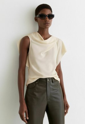 Kobieta w czarnych okularach przeciwsłonecznych w stylu cat-eye, bezrękawniku w kremowym kolorze z drapowaniem oraz w ciemnobrązowych, skórzanych spodniach z wysokim stanem, stojąca na tle białej ściany.