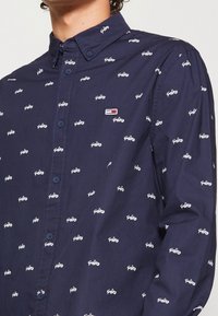 Mężczyzna w granatowej koszuli zapinanej na guziki, pokrytej małym białym napisem "Tommy Jeans" oraz małym prostokątnym logo w kolorach czerwonym, białym i granatowym.