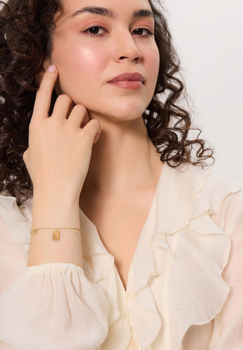 Złota bransoletka z prostokątnym charms'em i małymi akcentami serca, noszona na nagim nadgarstku, w zestawieniu z kremową bluzką z falbankami.