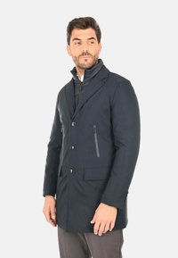 Cappotto impermeabile blu navy dal design lungo, con chiusura frontale a bottoni, due tasche e una tasca sul petto con cerniera; presenta un colletto alto e una texture liscia.