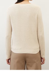 Pull beige côtelé à manches longues et col large, avec un tissage texturé et un ourlet légèrement incurvé. Porté avec un pantalon beige.