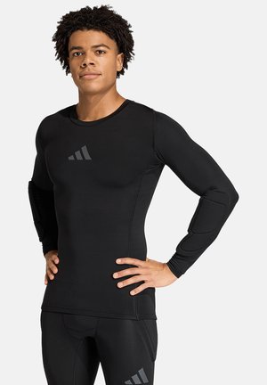 Jeune homme aux cheveux bouclés portant un haut de sport noir à manches longues et un pantalon avec des protections aux coudes, debout les mains sur les hanches.