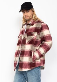 Hombre con cabello largo rubio usando una gorra negra, chaqueta a cuadros roja y beige, jeans de color azul claro, con una mano en el bolsillo de la chaqueta frente a un fondo blanco.